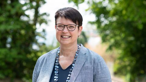 Regine Bresler Portrait von Regine Bresler, sie steht im Freien vor Bäumen und lächelt.