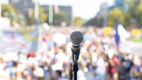 Ein Mikrofon steht in der Mitte des Bildes. Im Hintergrund sieht man unscharf eine Menschenmasse, die Schilder hochhalten und protestieren.