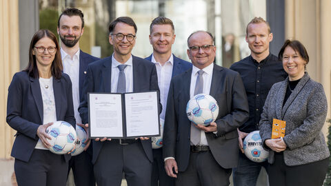 Justizminister Heinz steht mit weiteren Personen zusammen. Er hält ein Dokument in Händen, die anderen Personen halten Fußbälle.