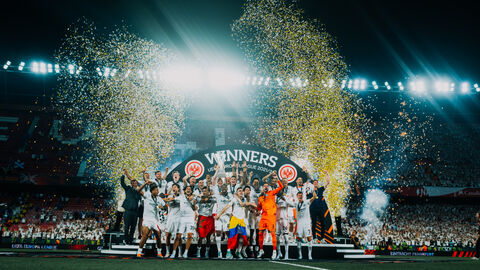 Fußballmannschaft steht in einem Stadion auf einer Bühne und feiert den Sieg. Sie reißen die Arme hoch und im Hintergrund fliegt goldenes Konfetti.