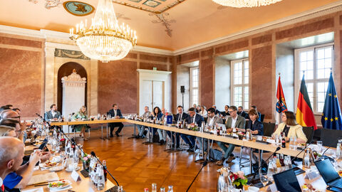 Eine große Konferenzrunde in einem historischen Saal mit zwei prächtigen Kronleuchtern, bei der die Teilnehmer an einer u-förmigen Tischreihe tagen.