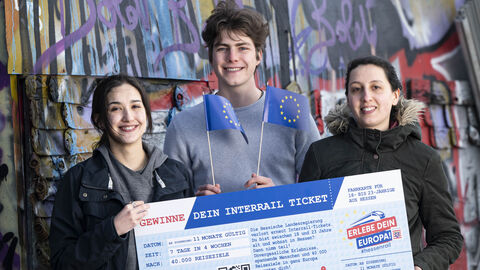 HessenRail-Auslosung Drei junge Menschen stehen vor einer Graffiti besprühten Wand mit einem "HessenRail"-Scheck in Händen. Die mittlere Person wedelt mit kleinen Europa-Flaggen