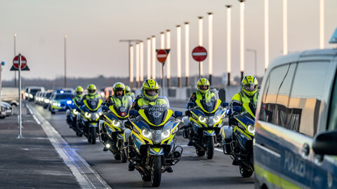 Mehrere Motorräder der Polizei sowie Autos und Busse der Polizei fahren hintereinander auf einer Straße.