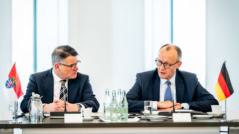 Zwei Männer sitzen an einem Tisch nebeneinander. An den Seiten des Tisches stehen die Hessische Flagge (links) und die Deutschland-Flagge (rechts).