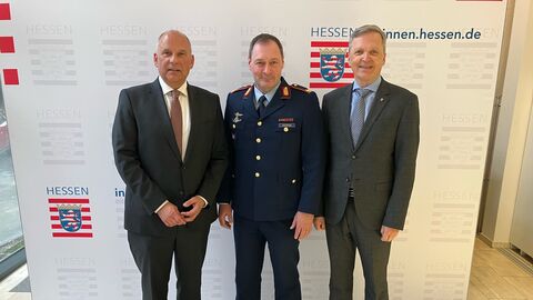 Drei Männer stehen nebeneinander vor einer Wand. Im Hintergrund steht eine Wand mit dem Wappen von Hessen.